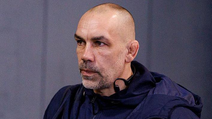 Alex Sanderson looking straight ahead with a grey wall behind him 