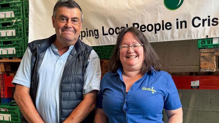 Mike and Maria smile at the camera while standing in a warehouse, part of a foodbank banner behind them. Mike is wearing a blue polo top and dark gilet while Maria wears a blue shirt with foodbank branding.