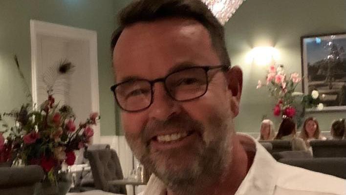 A man with short dark hair and black glasses smiles at the camera. He is wearing a white shirt and is sitting in a restaurant. There are flowers on a table behind him and diners on another table