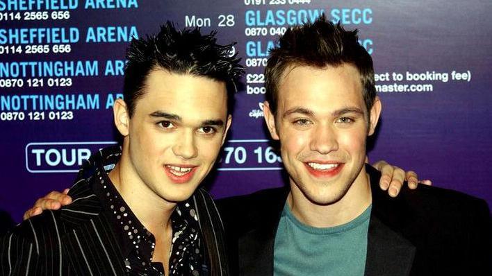 Two young men smile at the camera. Gareth Gates stands on the left wearing a striped suit jacket and spotty shirt and Will Young is on the right, wearing a green T-shirt and black suit jacket.