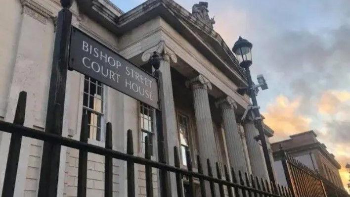 The outside of Bishop Street Court House in Londonderry. A street lamp and black gate is just outside a pillared building.