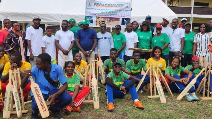 A group of around 30 people pose for a photo at a cricket event, with a Fecacricket sign in the background over the entrance to a large white gazebo. Eleven people sit or crouch on the floor amid cricket bats and stumps, with others standing behind them. Many are dressed in yellow and green sports tops