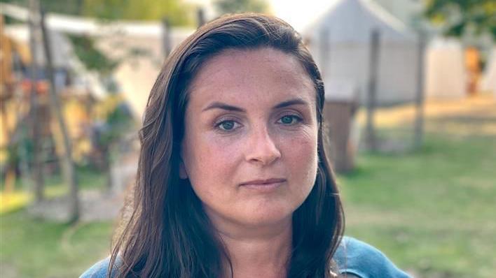 Lizzie Musson smiles at the camera. She has long brown hair which is parted to the side. She wears a blue cape. Behind her Anglo-Saxon recreated villages and scenes can be seen, 