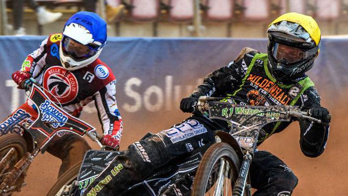 A Mildenhall Fen Tiger speedway rider is pictured mid-race making his way around a corner on a dirt track. He has a yellow helmet and black racing suit on with various designs and patterns on it. Another rider is behind him also making his way around a corner.