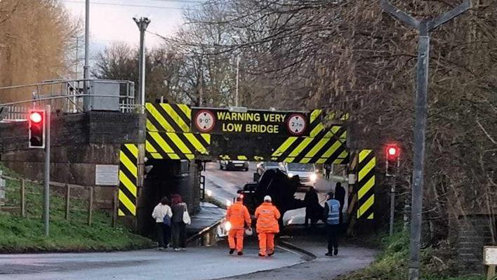 A bridge with yellow and black signs around it warning road users it is a 'very low bridge'. Underneath it a vehicle looks to be on its side. Two people in orange high visibility outfits are walking towards the blockage under the bridge. 