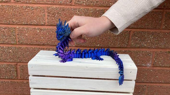 A hand holding a dark blue and purple 3D printed dragon on top of a light wooden box. There is a red brick wall in the background. 