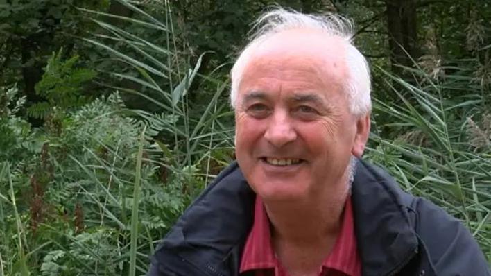 Prof Alastair Fitter wearing a red shirt with white stripes and a blue waterproof coat photographed in front of bracken and reeds at Askham Bog