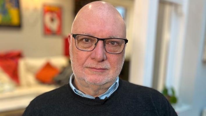 Mark Harrison looking directly at the camera. He is bald and has dark glasses and stubble. He is wearing a blue shirt and dark coloured sweater.