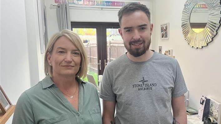 A woman in a green shirt and a man in a grey t-shirt, stood together in a hallway. Small pictures and a round mirror are hung on the walls.
