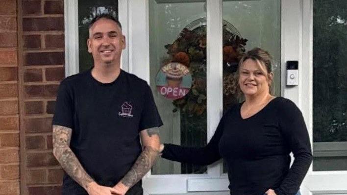A man and woman stand in front of a white porch. The man stands to our left and has short dark hair and both his arms are decorated with tattoos. He is wearing a black T-shirt with a cake logo on it. The woman is standing to our right. She has brown hair tied back and is wearing a long-sleeved black top. There is a small sign on the door which reads 'we are open'.