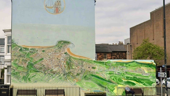A large mural painted on the side wall of a building in an urban street setting. The wall is painted a pale blue colour and features a detailed aerial-style map or landscape illustration across the lower half. The map appears to depict a coastal town, with a curved shoreline, sandy beaches, streets, buildings and large areas of green countryside. The painting extends beyond the edge of the wall on to a lower adjoining structure, creating a long panoramic view of the landscape.