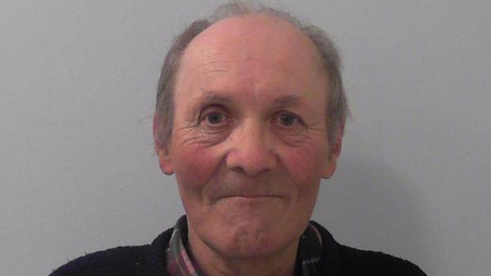 Phipps' mugshot, where he is smiling with thin pursed lips and rosy cheeks. He is balding and has grey hair. He is wearing a tartan shirt with a dark jumper or cardigan over it and is stood in front of a grey background.