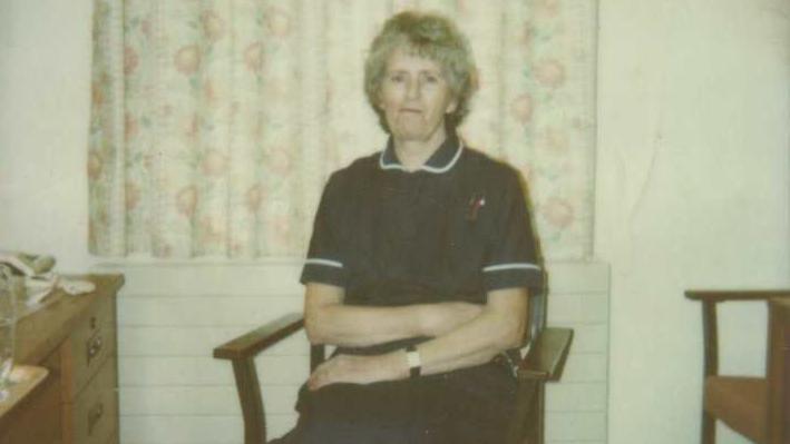 Nora sitting on a wooden chair, looking at the camera, in her navy and white nurses uniform.