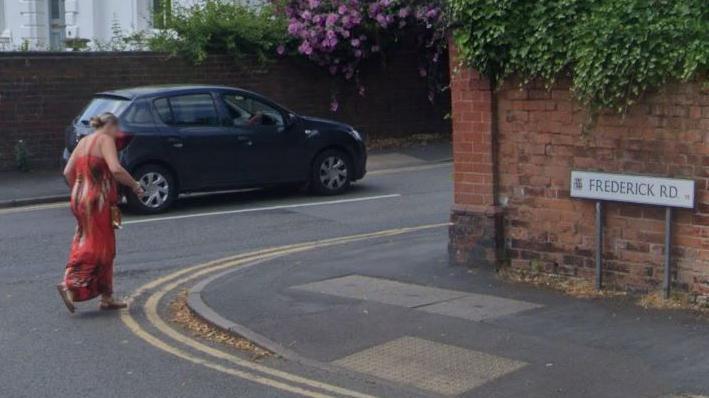 A woman dressed in a long red dress is crossing Frederick Road at its junction with St James Road, on which a blue car is being driven. The sign for Frederick Road is showing on the right of the shot. 