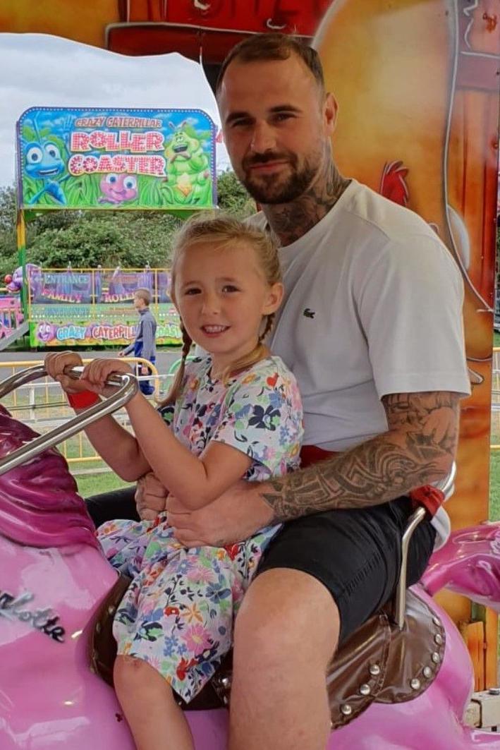 Tom Gaughan, who is wearing a light T-shirt and dark shorts, sits holding his young daughter, Kendall, who is wearing a floral dress, on a fairground ride.