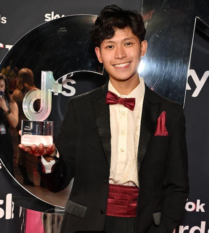 Gab Astorga holds a glass award up to the camera. He has black wavy hair and is wearing a black dinner suit with red bow tie and standing in front of a black branded screen.