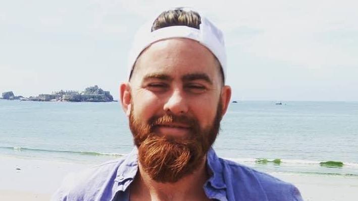 Frazer Irvine standing on a beach looking into a camera, wearing a white baseball cap