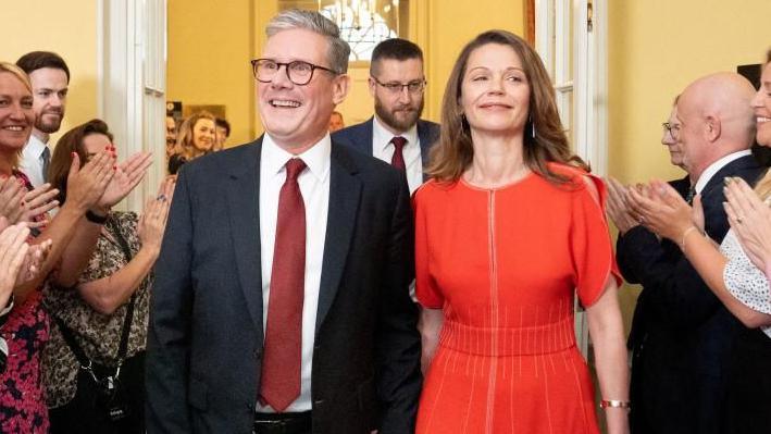 Sir Keir Starmer, wearing a suit, and his wife Victoria Starmer, wearing a red dress, are clapped in by staff as they enter his official London residence at No 10