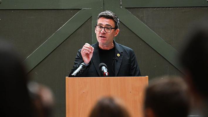Llun o Andy Burnham yn annerch cynulleidfa. Mae'n gwisgo dillad du ac yn sefyll y tu ôl i bodiwm.