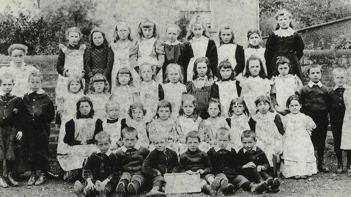 Pupils wearing traditional 20th Century clothes in a black and white photo. There are at least 30 children in the image, with a brick wall behind them.