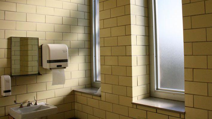Photo of yellow-tiled restroom. There is a sink with a mirror, soap dispenser and paper dispenser above. There are two windows on the wall beside the sink.