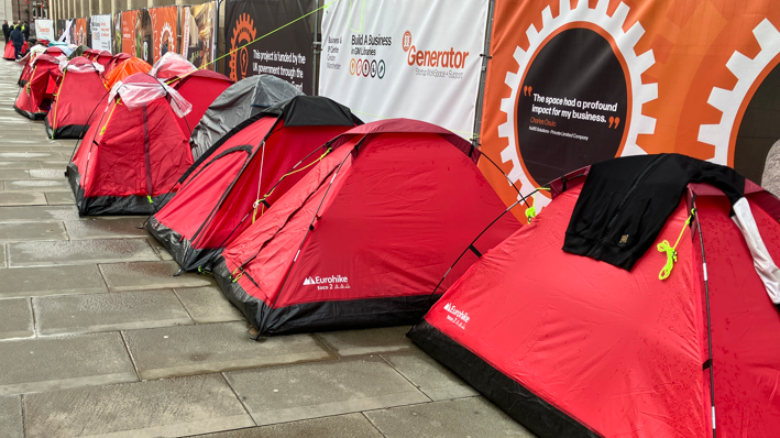 A line of red tents outside Manchester town hall, pitched on wet stone paving, under some fencing covered in advertising. 