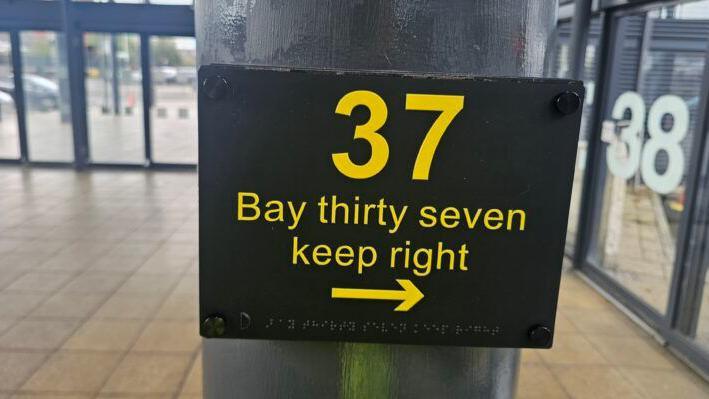 A black sign with yellow writing reads 'Bay thirty seven keep right' with a line of braille underneath it. You can see the bus station in the background of the photo.