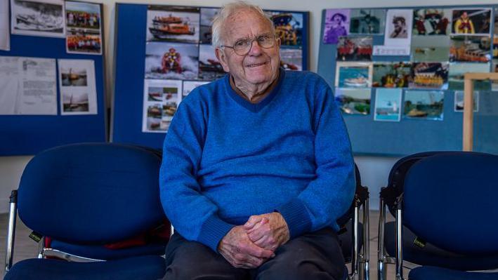 Bob McLaughlin sitting on a chair in front of photos which are part of an RNLI exhibition. He is smiling at the camera and has white hair, glasses and is wearing a bright blue jumper.