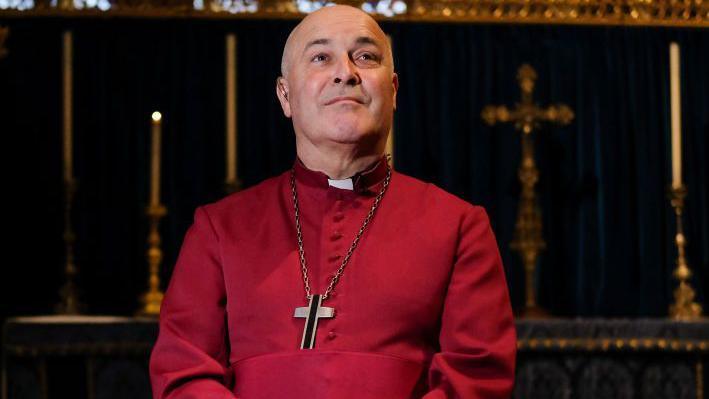 Stephen Cottrell in dark red clerical clothing with a Christian cross necklace on. He is bald and has light coloured eyes.