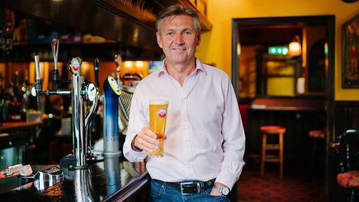 a man wearing jeans and a pale shirt holds a pint of lager and has one hand in his pocket next to a bar