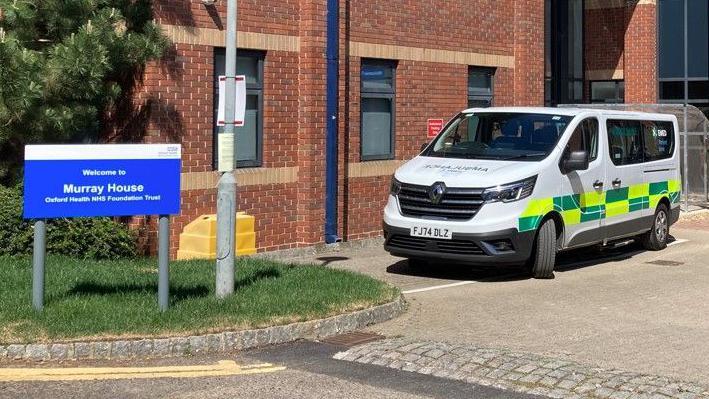 Outside a red brick building, a blue and white NHS sign reads "Welcome to Murray House". An ambulance is parked alongside the building.