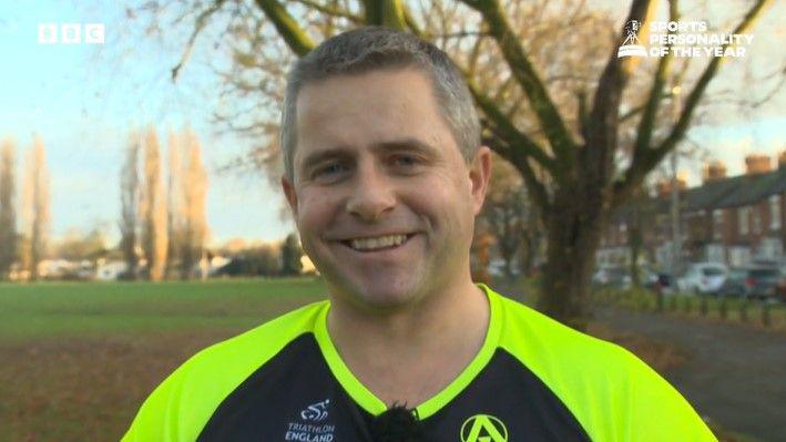 Paul McIntyre is wearing a high-visibility t-shirt which is black with fluorescent yellow shoulders and collar. He is stood on a playing field, with trees and an adjacent row of houses visible behind him.