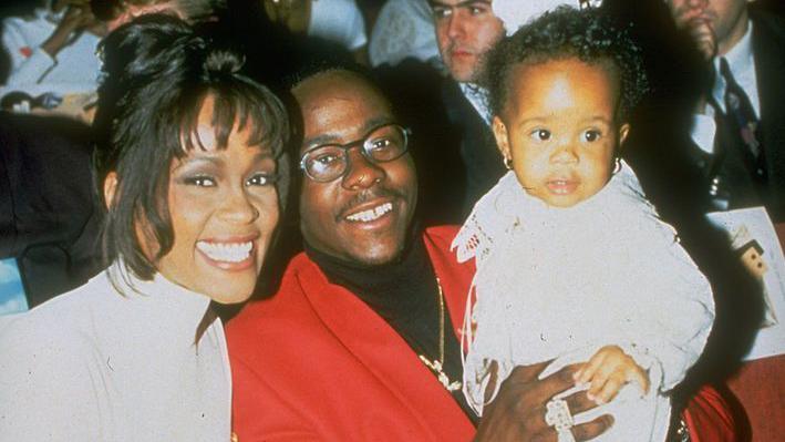 Whitney Houston sitting with her singer husband Bobby Brown, who is holding their infant daughter Bobbi. All are smiling. Houston has her hair up and is wearing a white turtle neck top, Brown is wearing a red blazer and black turtle neck with a gold chain around his neck, Bobbi has pierced ears and is all in white.