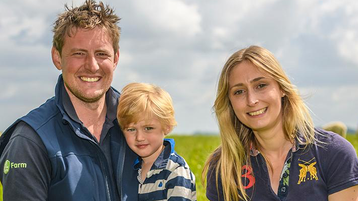 Sam and Samantha Edwards and their young son smiling for the camera. They are wearing navy blue t-shirts and dark jeans. Behind them, out of focus, are some sheep.