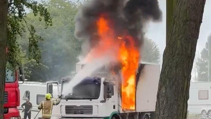 A firefighter points a hose at a lorry trailer which is alight at the front section