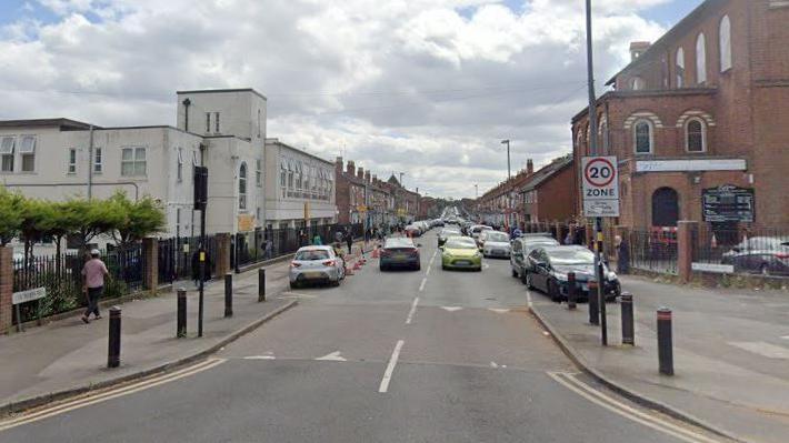 A built-up street with a 20mph zone sign and buildings on either side. Cars drive down on both sides and there are also cars parked on the side.