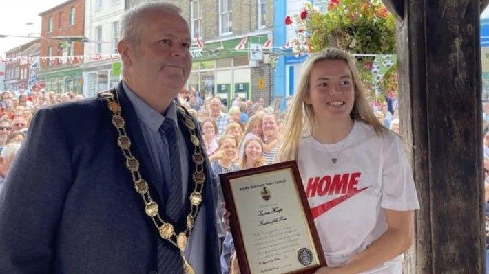 A male mayor wearing a suit and ceremonial chains hands a certificate to a blonde woman wearing a white t-shirt with a large crowd gathered behind them.