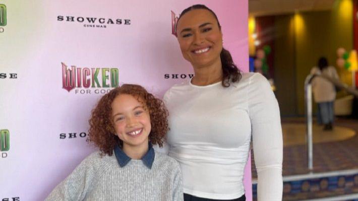 Esme posing with her mother Shareen, who has her arm around her and is wearing a white top and black trousers.
