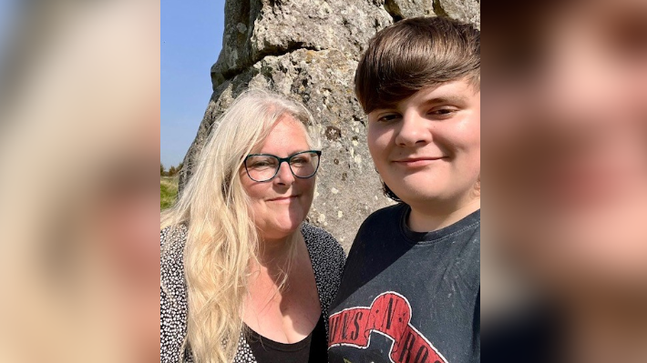 Dylan with his mum stood in front of a large rock in the countryside smiling.
Dylan is wearing a black t-shirt and his mum with wearing a black top and glasses.