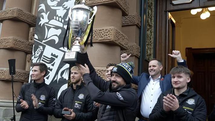 The Ipswich Witches team raise their trophy while standing on the tops outside a town hall. They clap and cheer and some raise their arms in praise.