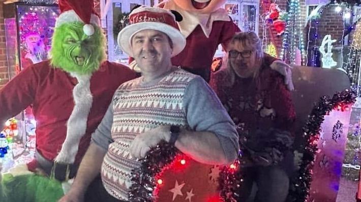 The couple are sat next to the Grinch on a large sleigh outside their home. Mr Greenhouse wears a Christmas jumper with a stripey design and a red hat with white rim. The Grinch is pulling a face while wearing a Santa outfit. Mrs Greenhouse has blonde hair to her shoulders and wears a red jumper with white reindeer on it.