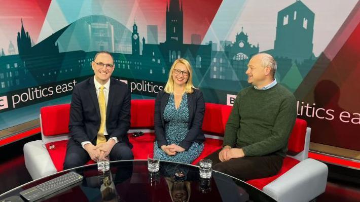 Ian Sollom, Jess Asato and Jerome Mayhew sit in a row on a red sofa in a TV studio. Sollom is wearing a suit, Asato is wearing a blue patterned dress and navy jacket, and Mayhew is wearing a green jumper, blue shirt and dark trousers. They are all smiling. There is a desk in front of them with a computer keyboard and three glasses of water. 