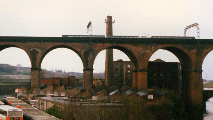 An historic, slightly grainy, photo of the viaduct, with arches standing imposingly in front of a mill building, with homes and a busy station seen below on an overcast day. A train runs along the viaduct.