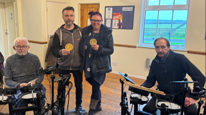 Phil Jones is standing next to a woman holding up a gold medal. Next to them are two participants sitting at electric drum kits.