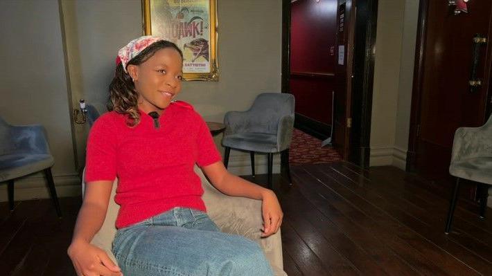 A woman wearing a bright pink jumper sat in a chair. She is wearing a white and pink flowery headscarf and blue jeans. 