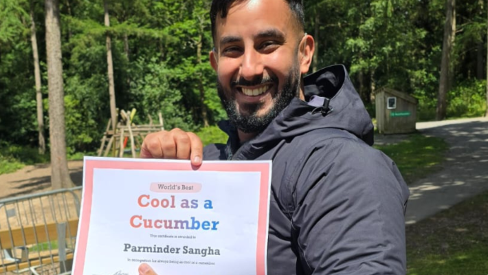 Parminder Sangha outside in a green space wearing a black hooded jacket holding a piece of paper that says "Cool as a Cucumber, Parminder Sangha"