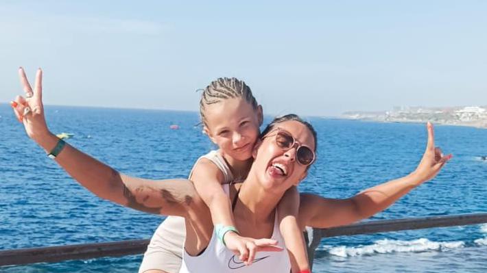 Mum in sunglasses has tongue out, smiling with daughter on her shoulders. Their arms are out making peace signs, there is a body of water behind them and blue sky. 