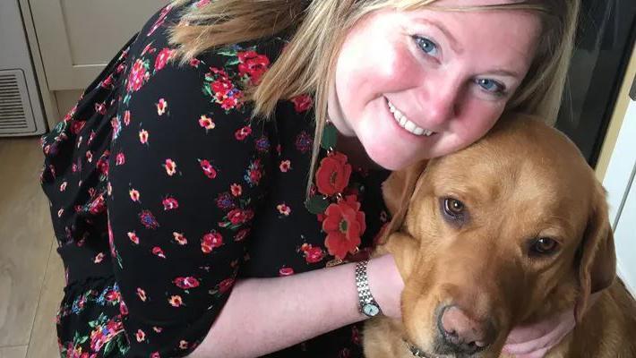 Gemma Fairhurst, smiling with blonde hair, wears a black dress with a flowery pattern. She is crouched down with her face cheek-to-cheek with guide dog Ian.