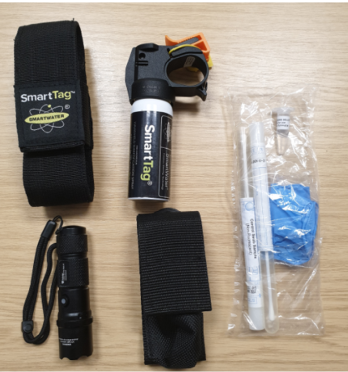 Photograph shows the Smart Tag equipment laid out on a table. There is a plastic clear bag including two swabs and a solution, a black cylinder hand held device with a black strap, along with the spray bottle which reads "smart tag" and a "smart tag" case.