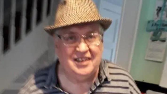 A smiling older man looks to the camera. He is wearing a hat, glasses and an open-neck shirt. He looks happy.